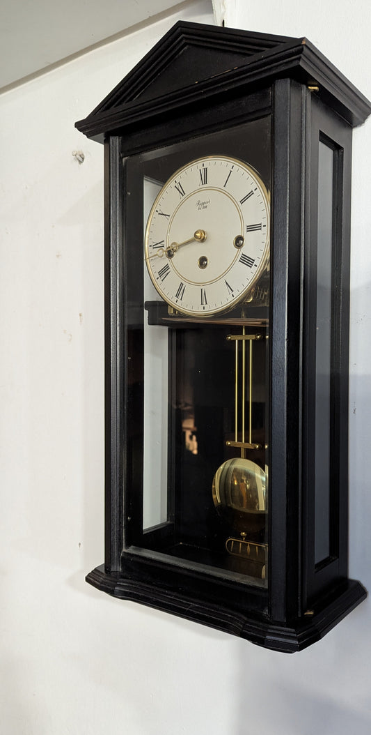 A mechanical Westminster wall clock by Rapport in ebony black with glass sides and front, showing the clock face and internal mechanism.