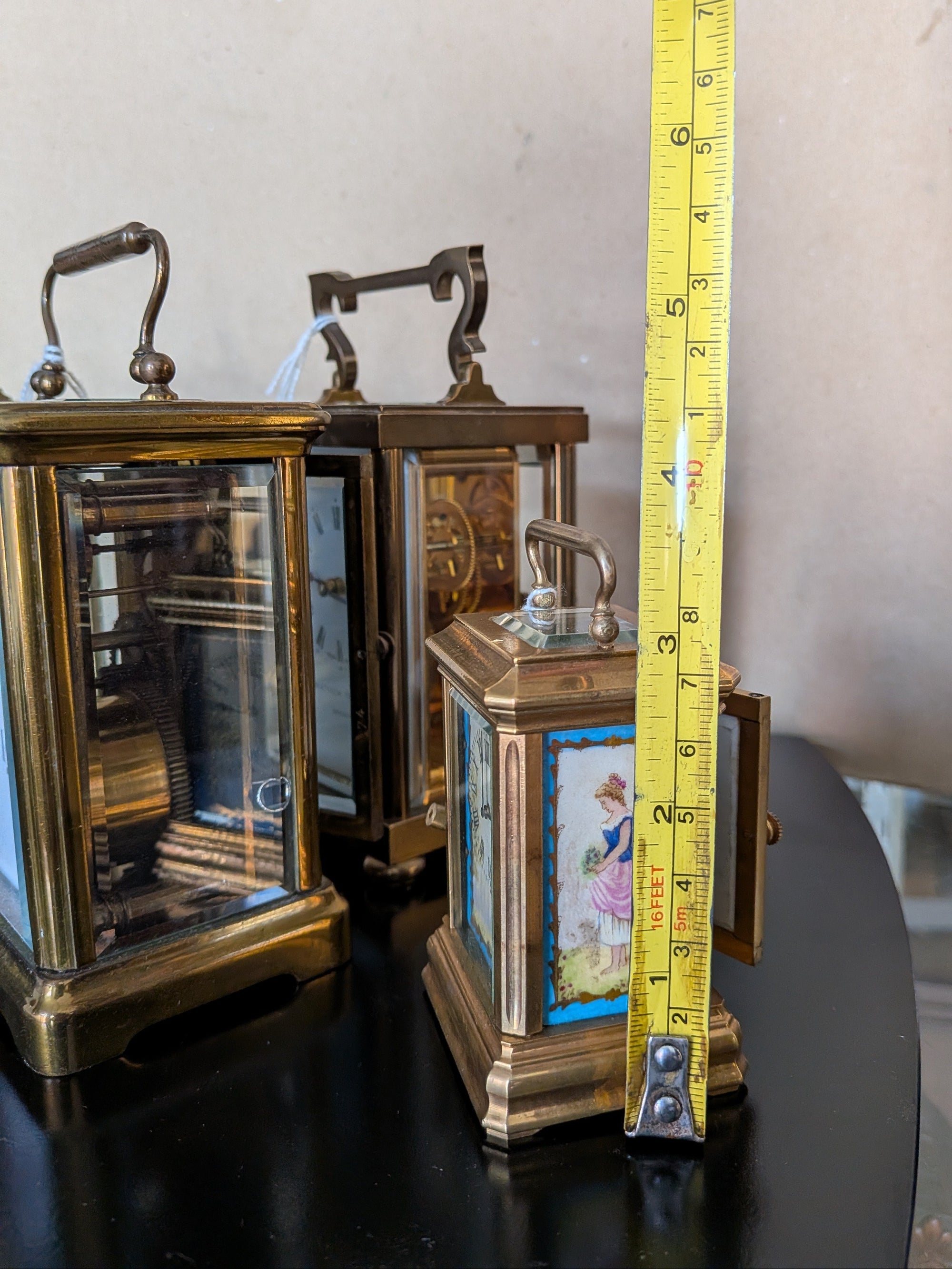 Three brass clocks with a measuring tape for scale on a reflective surface.